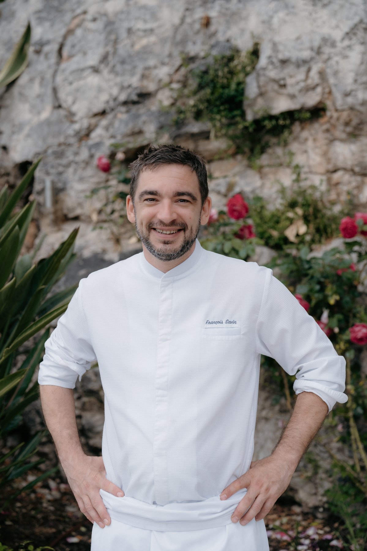 Chef François Savin in his whites, outdoor portrait with a Provençal stone wall and red roses behind.