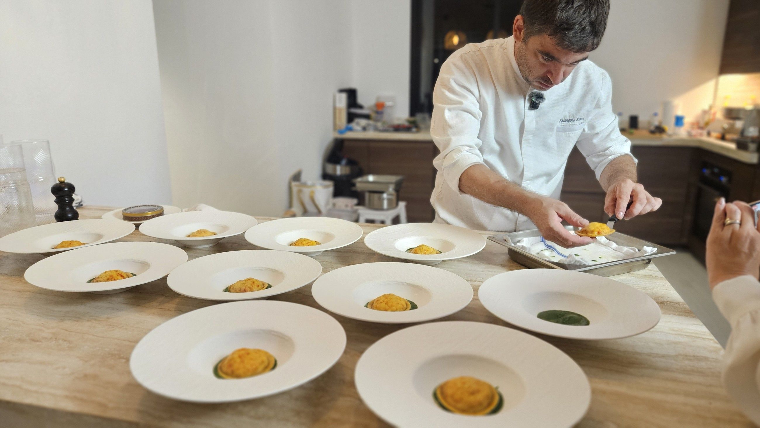 Chef François Savin in his whites, plating a series of dishes on white bowls at a villa kitchen counter.