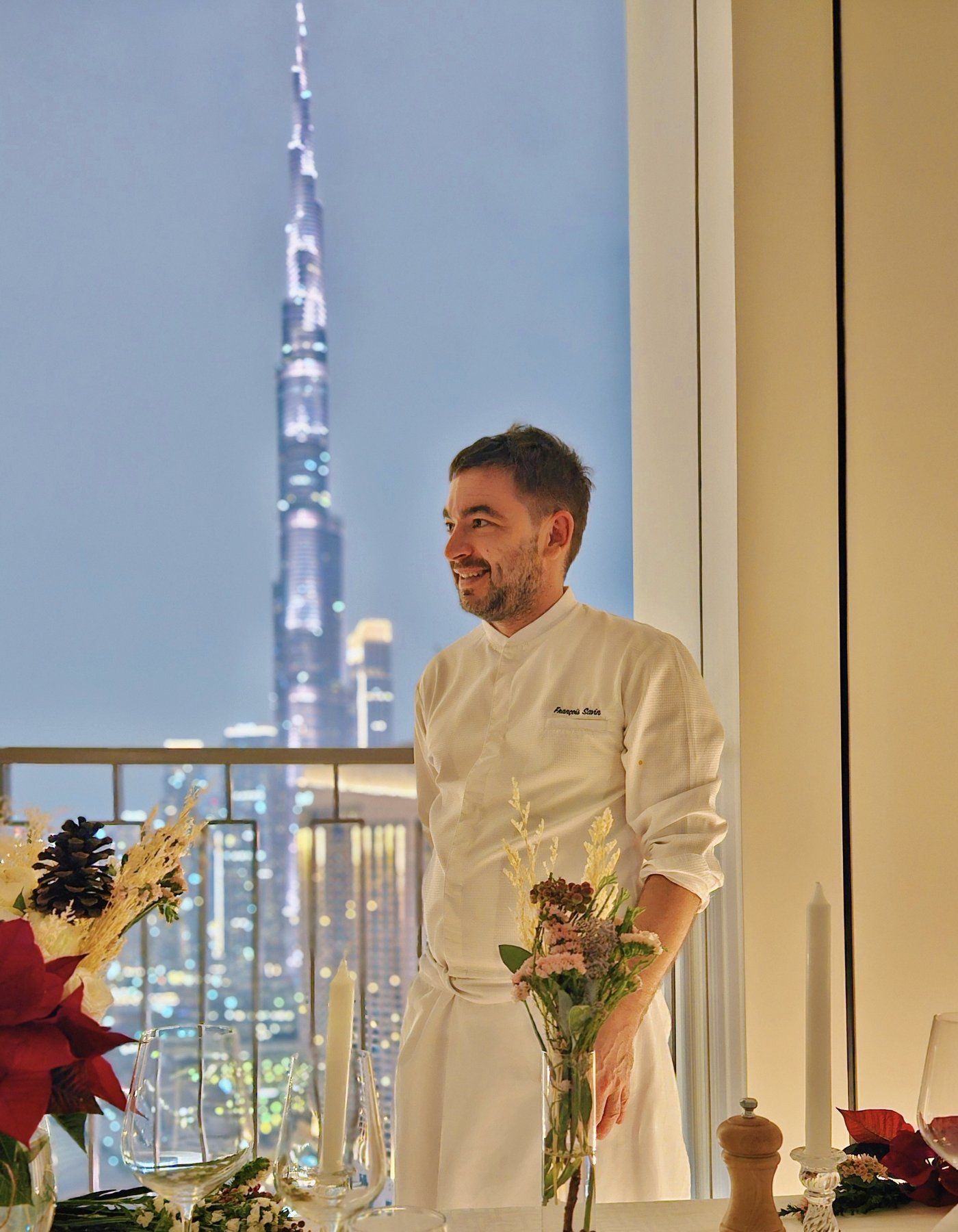 Chef François Savin in his whites on a Dubai high-rise terrace, table dressed with white linen and red blooms, skyline behind.