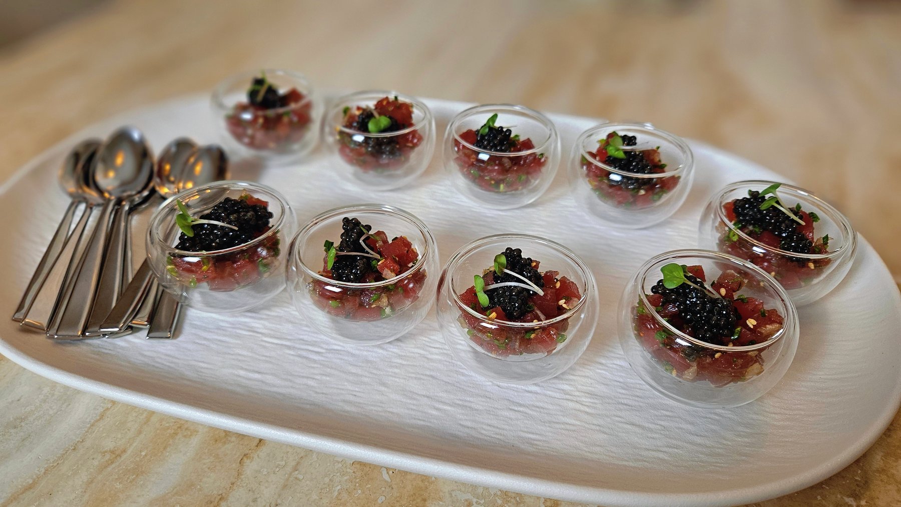 Tuna tartare bites topped with caviar, served in glass spoons on a white tray.